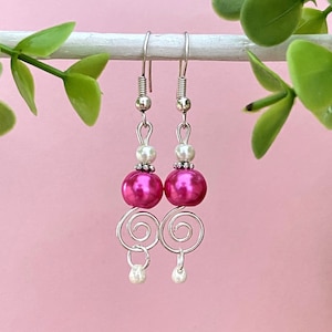Puede incluir: Pendientes de perlas rosas y blancas con espirales de alambre plateado. Los pendientes cuelgan de una superficie blanca.