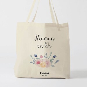 May include: A beige tote bag with the text "Maman en Or" and a floral design in shades of pink, yellow, and blue. The bag also has the text "L'atelier des Amis" at the bottom.