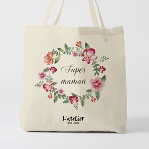 Può includere: Una borsa tote in tela bianca con un design a corona di fiori e il testo "Super maman" in corsivo nero. La parte inferiore della borsa ha il testo "L'atelier des amis" in nero.