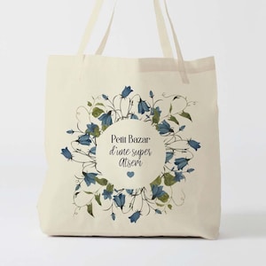 May include: Cream-colored tote bag with a floral design. The design features blue and green flowers arranged in a circular pattern around a central text. The text reads "Petit Bazar d'une super atsem".