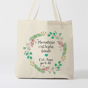 Peut inclure: Un sac fourre-tout en toile blanche avec un motif de couronne florale verte. Le texte à l'intérieur de la couronne dit "Ma maitresse c'est la plus Geniale C'est Anna qui le dit".