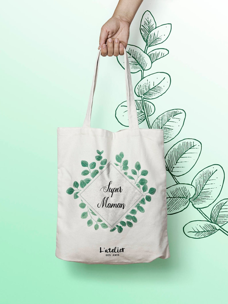 May include: White tote bag with a green floral design and the text "Super Maman" in black. The bag also has the text "L'atelier des amis" in black.