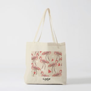 May include: A beige tote bag with a pink flamingo pattern and the text "L'atelier des amis" printed on the front.