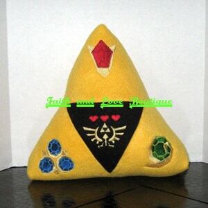 May include: A yellow triangular pillow with a black triangular patch in the center. The patch has a stylized design with three hearts and a winged symbol. The pillow has three embroidered gems, one red, one green, and one blue.