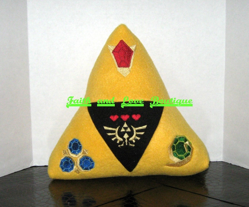 May include: A yellow triangular pillow with a black triangular patch in the center. The patch has a stylized design with three hearts and a winged symbol. The pillow has three embroidered gems, one red, one green, and one blue.