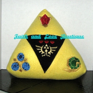 May include: A yellow triangular pillow with embroidered images of the Triforce, a red heart, and three blue and green gems. The pillow is decorated with gold stitching.