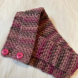 Puede incluir: Un calentador de cuello de ganchillo en tonos rosa, marrón y gris con dos botones rosas.