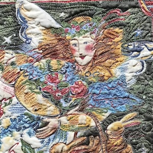 Puede incluir: Una obra de arte bordada de carácter fantástico que representa a un ángel alado con una corona de flores, sosteniendo una cesta con un conejo en su interior. El ángel está rodeado de flores y hojas, y el fondo es de color verde oscuro.