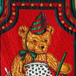 May include: A red quilted oven mitt with a green border features a festive Christmas scene with a teddy bear wearing a red and green hat, holding a wooden spoon and a bowl of red jam. The scene includes holly, gingerbread cookies, and a book titled "Christmas Pudding Recipes".