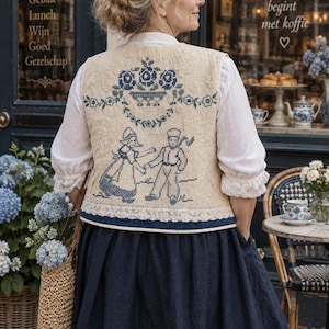 May include: A cream-colored vest featuring blue cross-stitch embroidery of a floral basket and two figures in traditional attire. The garment includes a white lace trim along the bottom edge and is worn over a white blouse and a dark blue skirt.