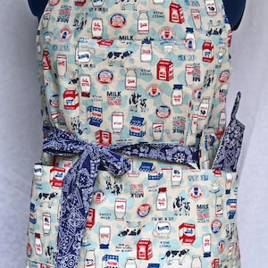 May include: A blue and white apron with a milk carton pattern. The apron has a large pocket and a tie at the waist. The apron is decorated with images of milk cartons, cows, and the words "Milk Crown" and "Give Me Some Milk".