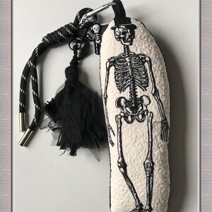 May include: A cream-colored, textured keychain featuring a black skeleton illustration wearing a top hat. The keychain has a black and white cord, a black tassel, and a small skeleton charm. The design is gothic and spooky.
