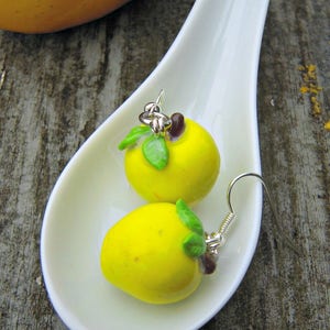 Peut inclure: Une paire de boucles d'oreilles en forme de pomme jaune avec des feuilles vertes et des accents marrons. Les boucles d'oreilles sont sur un plat blanc en forme de cuillère. Le texte "Le temps des pommes" est visible en bas de l'image.