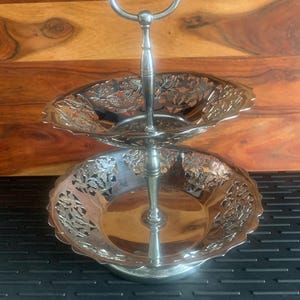 May include: A two-tiered silver-colored serving tray with a handle. The trays have a scalloped edge and a decorative floral design. The handle is a loop at the top. The tray is on a black surface with a wooden background.