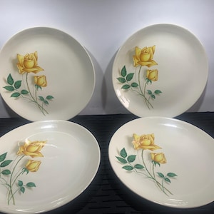 May include: Four white ceramic plates with a yellow rose design. Each plate features a yellow rose with green leaves and stems. Two plates are round, and two are oval-shaped, suitable for serving food.