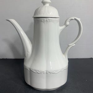 May include: A white porcelain coffee pot with a lid and a curved handle. The pot has a long spout and decorative embossed detailing around the base and on the lid. The design is classic and elegant.