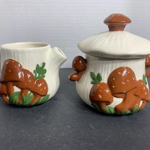 May include: Cream-colored ceramic creamer and sugar bowl set. The set is decorated with brown mushrooms with white spots and green leaves. The sugar bowl has a lid with a mushroom-shaped handle.