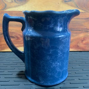 May include: A blue ceramic pitcher with a handle and a spout. The pitcher has a marbled effect and a scalloped rim. The handle is curved and attached to the side of the pitcher. The pitcher is sitting on a dark surface.