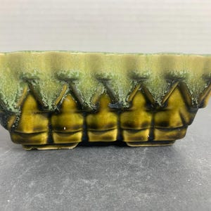 May include: Rectangular ceramic planter with a textured, scalloped rim. The planter features a geometric design with a blend of green and yellow-brown hues. The planter is suitable for small plants or succulents.