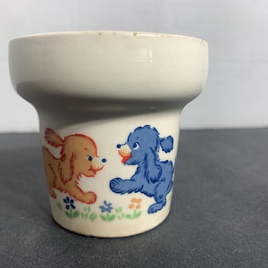 May include: A cream-colored ceramic cup featuring a graphic illustration of two small dogs, one orange and one blue, playing among small green and blue flowers. The cup has a wide, flared rim and a tapered body with a smooth, glossy finish.