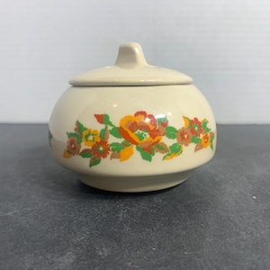 May include: A cream-colored ceramic sugar bowl with a lid. The bowl is decorated with a floral pattern in shades of orange, yellow, and green. The lid has a small handle. The bowl is round and sits on a flat base.