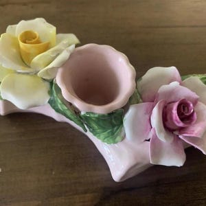 May include: A ceramic floral centerpiece featuring a central pink vase flanked by two roses. One rose is yellow and white, the other is pink and white. Green leaves accent the flowers and vase, all on a light pink base.