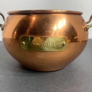 May include: A copper potpourri bowl with brass handles and a brass nameplate. The bowl has a rounded shape and a polished finish, reflecting light. The nameplate reads "POTPOURRI".