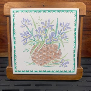 May include: A square ceramic tile featuring a floral design of purple and yellow irises in a woven basket. The tile is set in a wooden holder. The tile has a green border with a white lace-like trim.