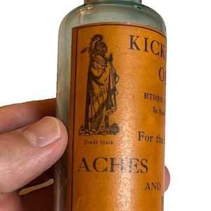 Puede incluir: Una botella vintage de Kickapoo Indian con una etiqueta naranja y texto negro. La etiqueta presenta una ilustración de una figura nativa americana y anuncia un producto para dolores. La botella es de vidrio transparente.