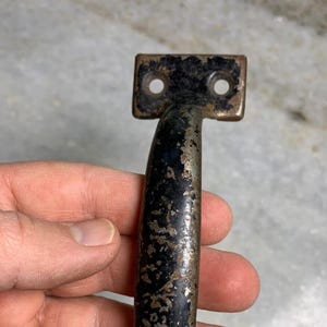 Könnte beinhalten: Ein schwarzer Metalltürgriff mit abgerundeter Form und zwei quadratischen Platten mit Löchern für Schrauben. Der Griff hat ein abgenutztes, gealtertes Aussehen, wobei etwas Rost durch die schwarze Farbe sichtbar ist.