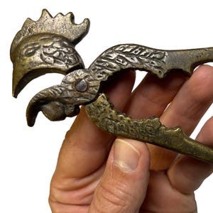 May include: A metallic nutcracker shaped like a rooster with a weathered bronze finish. The tool features a hinged design with textured detailing on the bird's head and neck, and two long handles that extend to form the body and tail of the rooster.