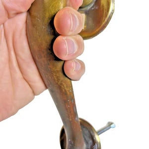 May include: Vintage brass door handle with a curved design. The handle has a tarnished finish, showing signs of age and use. The handle is held in a hand, showcasing its size and shape. Includes a mounting screw.