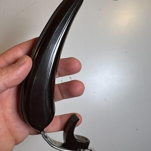 Puede incluir: Un tirador curvo de color marrón oscuro con un diseño acanalado, sujeto a un soporte metálico plateado. El tirador se sostiene en una mano sobre un fondo blanco. El tirador parece ser una pieza de mobiliario vintage o antiguo.