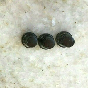 Puede incluir: Tres perillas negras con borde estriado y parte superior redondeada dispuestas en línea horizontal. Las perillas parecen ser de metal y están sobre una superficie clara y moteada.