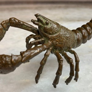 Puede incluir: Una escultura de bronce de un cangrejo de río con garras detalladas y una cola segmentada. La escultura está en una pose de caminar con las garras levantadas.