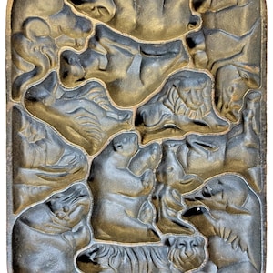 May include: A cast iron waffle iron with an animal theme. The rectangular iron features detailed relief images of various animals, including a tiger, bear, and lion. The iron has a handle on each end and is finished in a dark, metallic colour.