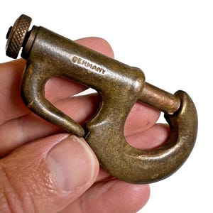 May include: A vintage brass-toned metal hook fastener with a textured, cylindrical screw-top mechanism. The curved, C-shaped body features a prominent stamped inscription that reads GERMANY. The metal surface shows a weathered, aged patina.