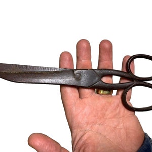 May include: Antique, heavy-duty shears with a dark, aged metal finish. The blades are wide and straight, with a textured cutting edge. The handles have large, round finger holes. A vintage tool for various cutting tasks.