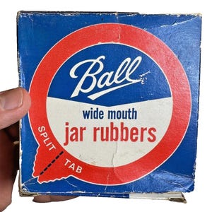Op de afbeelding: Een vintage blauwe doos met een rood en wit cirkelvormig ontwerp met het woord "Ball" en de tekst "wide mouth jar rubbers". De doos heeft een split-tab voor eenvoudig openen.