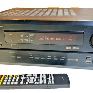 Pode incluir: Um receptor Denon AVR-2803 preto com controle remoto. O visor digital mostra "-A2 FM 89.10MHz -73". O controle remoto tem vários botões coloridos.