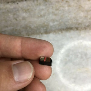 May include: A small, rectangular black object with a red and gold accent is held between fingers. The object appears to be a miniature electronic component or a small decorative piece. The background is a light-colored, possibly marble, surface.