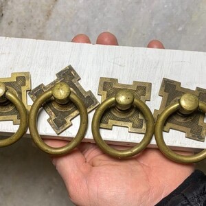 May include: Six antique brass cabinet knobs with square plates and ring pulls. The knobs are arranged in a row on a white surface.
