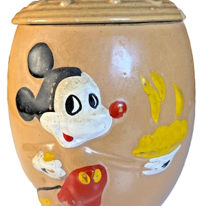 Op de afbeelding: Vintage Mickey Mouse koekjespot. De pot is bruin met een deksel en een knop. Mickey Mouse is afgebeeld op de voorkant in zwart, wit, rood en geel. De pot heeft een ronde vorm.