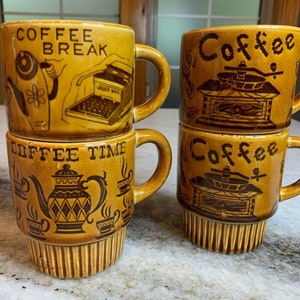 May include: Set of four brown ceramic coffee mugs with different coffee-themed designs. The mugs are stacked in pairs. One pair has the words "Coffee Break" and a cartoon of a person making coffee. The other pair has the words "Coffee Time" and a cartoon of a teapot. The mugs are all decorated with brown and black designs.