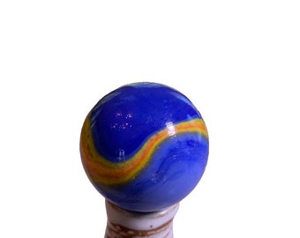May include: A blue glass marble with a yellow and orange swirl design. The marble is sitting on a white ceramic pedestal.