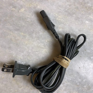 Può includere: Un cavo elettrico nero con una spina americana a un'estremità e una spina più piccola all'altra estremità. Il cavo è avvolto e fissato con un elastico.