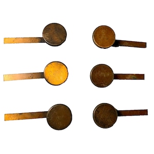 May include: Six antique brass-coloured metal knobs with a round shape and a small, flat, rectangular handle attached to the side. The knobs are arranged in two rows of three.
