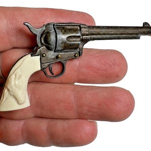 Puede incluir: Un revólver de juguete en miniatura con un acabado gris metálico desgastado, un mango de color crema con un diseño de cabeza de animal en relieve y una punta de seguridad naranja brillante en el cañón, presentado a pequeña escala.