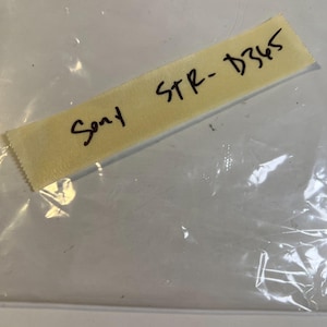 Puede incluir: Una etiqueta amarilla con el texto escrito a mano "Sony STR-D365" en tinta negra. La etiqueta está colocada dentro de una bolsa de plástico transparente, con los pliegues y arrugas de la bolsa visibles.