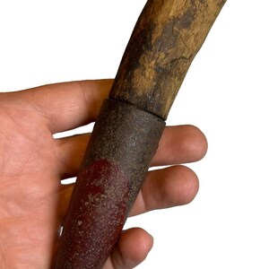 May include: A rustic, hand-carved wooden knife with a dark, weathered handle and a reddish-brown blade. The knife features a metal ferrule where the handle meets the blade. The blade has a pointed tip.